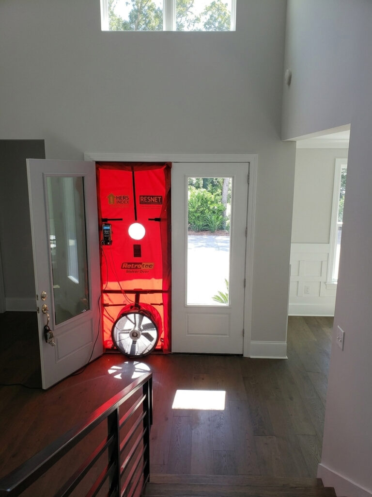 A photograph taken in aTampa Bay home from a staircase looks down on a red fabric blower door system installed in the frame of a modern front entry door. The system includes a large, powerful fan at the bottom, an upper viewing port, and a digital pressure gauge mounted to the left with various cables connected. Clear text and logos are visible on the red fabric, including "HERS INDEX," "RESNET," and "RETROTEC Blower Door." The other half of the doorway is a solid white paneled door with a full-length glass panel and brushed bronze hardware with keys. The floor is dark wood planks with a precise square patch of bright sunlight shining on it. To the right, an adjacent room with a white door and detailed wall paneling can be seen through an arched opening. The staircase in the foreground has a metal slatted balustrade. The interior walls are painted a soft grey. Natural light streams from high windows above the main entry door, which are partially obscured by the frame, and through the glass doors.